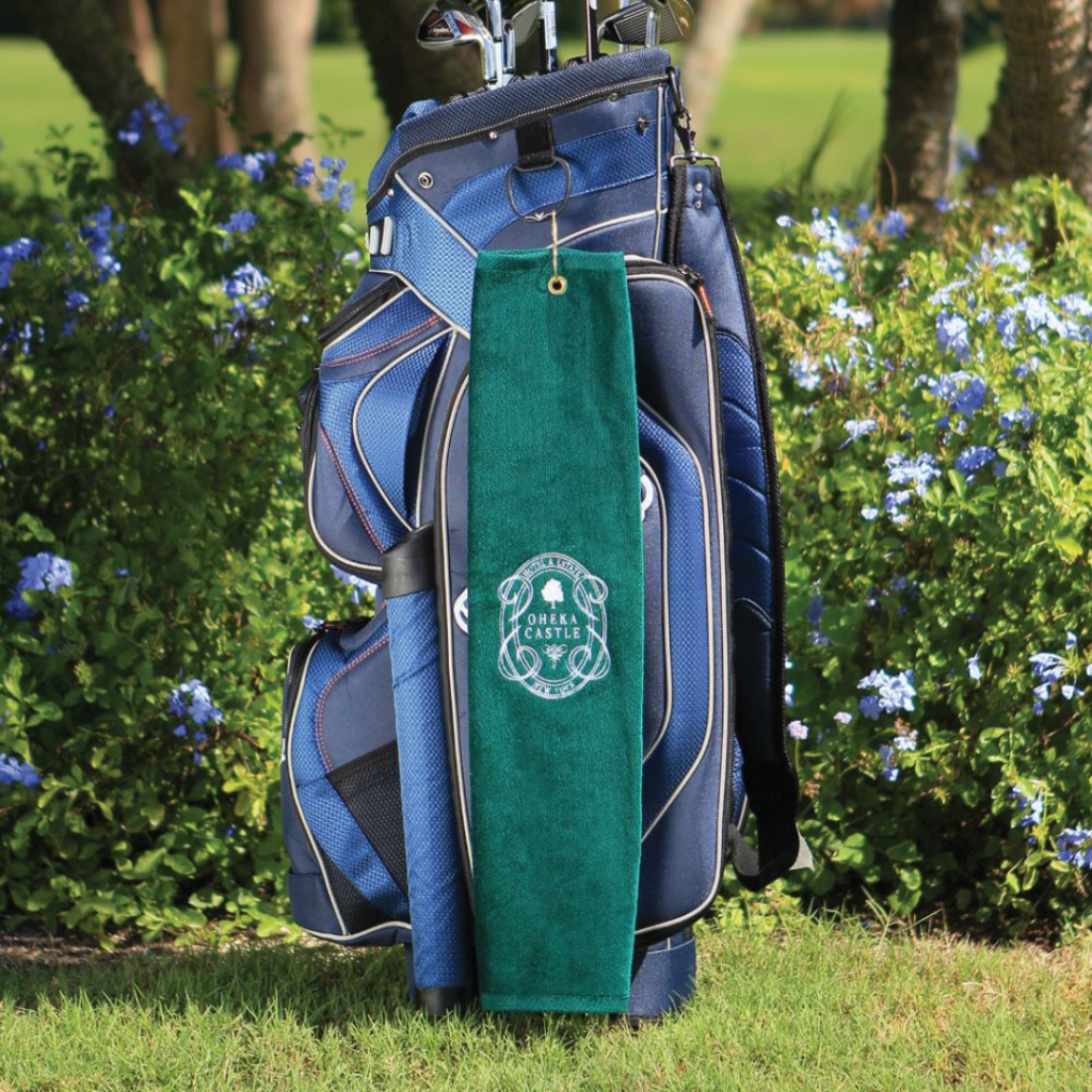 A blue golf bag sits on grass. Attached to it via a grommet is a custom branded green golf towel with logo on it