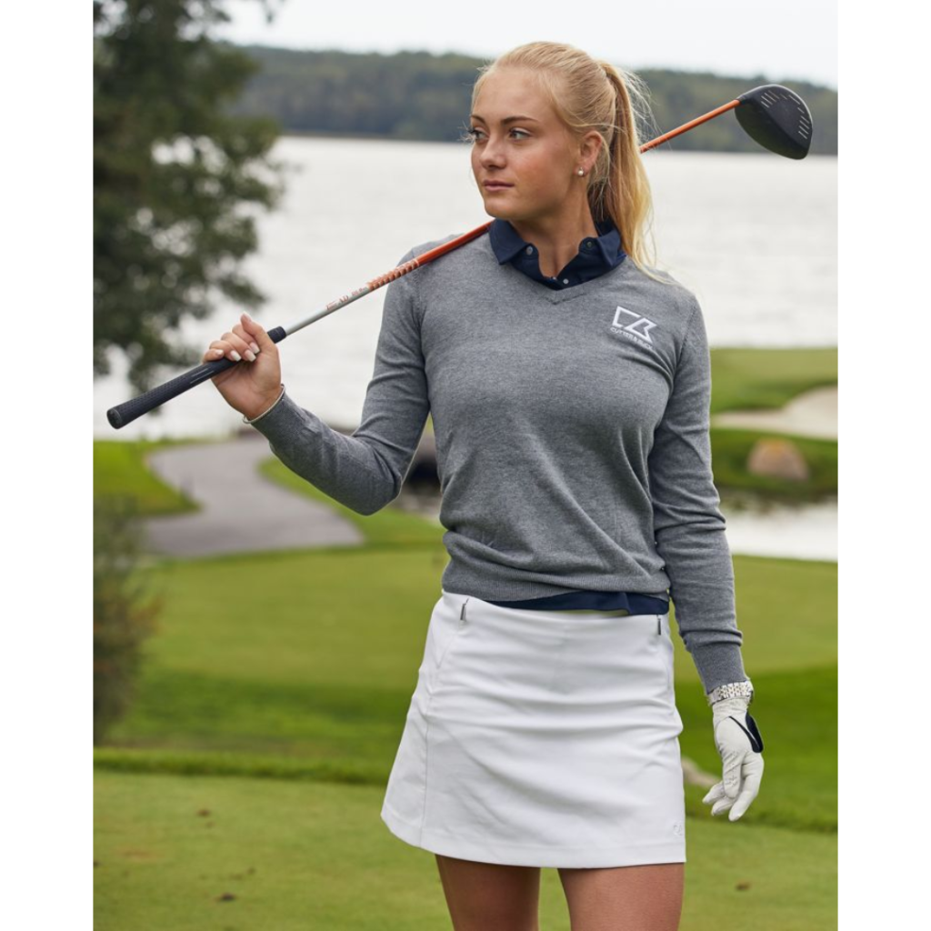 A blonde woman stands on a golf course with a golf club over her shoulder. She's wearing a grey custom branded Cutter and Buck shirt and white golf skirt